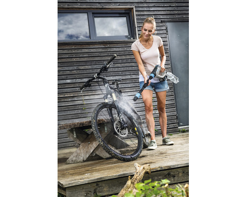 Frau reinigt Fahrrad mit einem Akku-Hochdruckreiniger vor einem Haus.