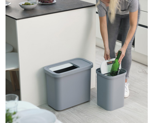 Deux poubelles grises dans une cuisine, une personne y jette une bouteille.
