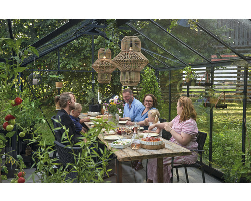 Scène dans une serre avec une famille assise à une table en bois, entourée de plantes et de deux suspensions.