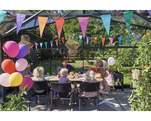 Une famille fête un anniversaire dans une serre avec une table, des chaises, des ballons et des guirlandes.