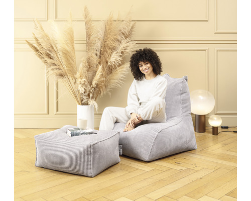 Femme assise sur un pouf en velours côtelé gris avec un tabouret assorti dans une pièce lumineuse.