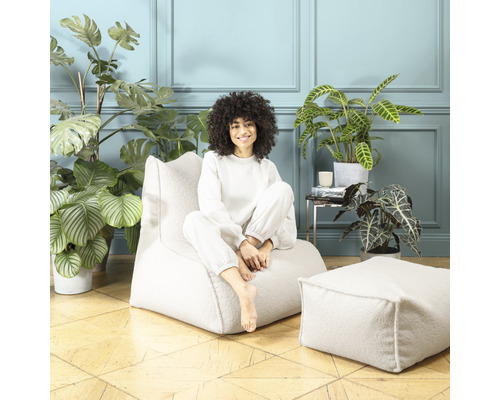 Femme assise sur un pouf blanc avec repose-pieds assorti, entourée de plantes d''intérieur dans une pièce lumineuse.