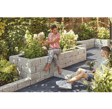 Jardin avec parterre surélevé en pierre, planté de fleurs, sur lequel une femme est assise, tandis qu''un homme est assis par terre à côté