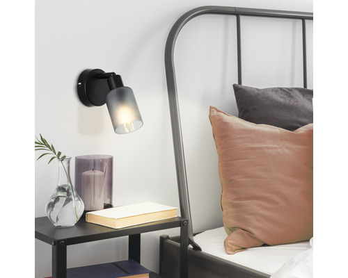 Wandleuchte aus Metall mit einem Lampenschirm aus Glas über einem Nachttisch mit Dekoration.