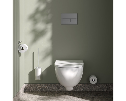 Wandhängendes WC mit Betätigungsplatte, Toilettenpapierhalter und Toilettenbürste in einem Badezimmer.