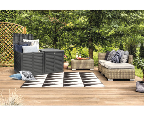 Gartenszene mit grauem Aufbewahrungsschrank, Rattansofa und schwarz-weißem Teppich auf einer Holzterrasse.
