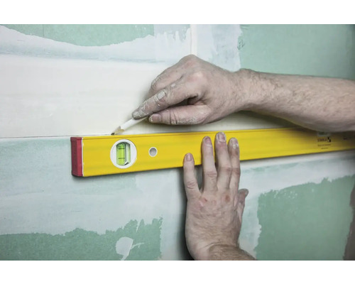 Eine Wasserwaage wird an einer Wand mit Gips befestigt, eine Person markiert mit einem Bleistift eine Linie.