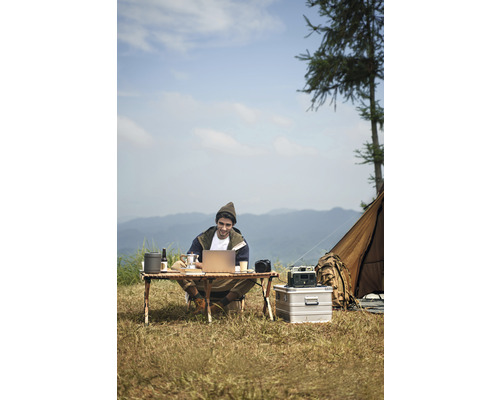 Mann sitzt am Campingtisch und arbeitet am Laptop im Freien mit Zelt im Hintergrund.