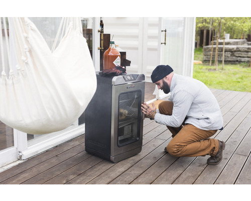 Un homme vérifie un fumoir à l''extérieur, à côté duquel se trouvent un hamac et une bouteille de sauce.