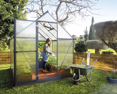 Gewächshaus mit geöffneter Tür in einem Garten, Frau giesst Pflanzen.