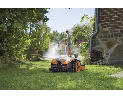 Robot tondeuse sur gazon, à l''arrière-plan, une personne nettoie une terrasse avec un nettoyeur haute pression