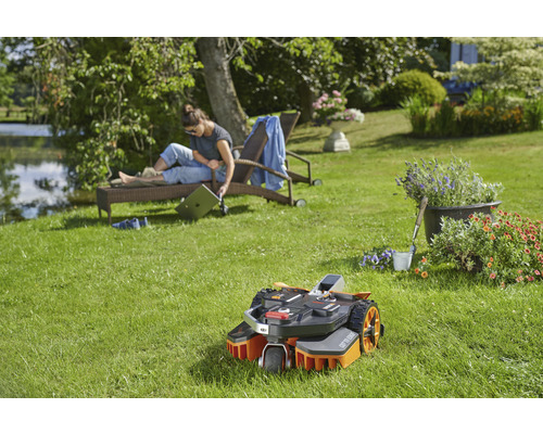 Szene mit einem Rasenmähroboter auf einer grünen Rasenfläche, während eine Frau sich auf einer Sonnenliege entspannt