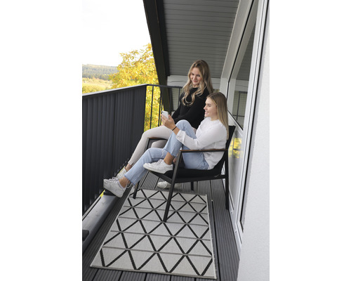 Szene auf einem Balkon mit zwei Frauen, Stühlen und einem Teppich mit geometrischem Muster.