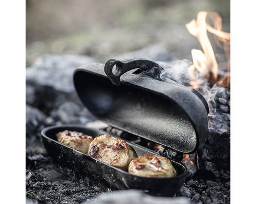 Offene Camping-Grillpfanne mit drei Kartoffeln am Lagerfeuer.