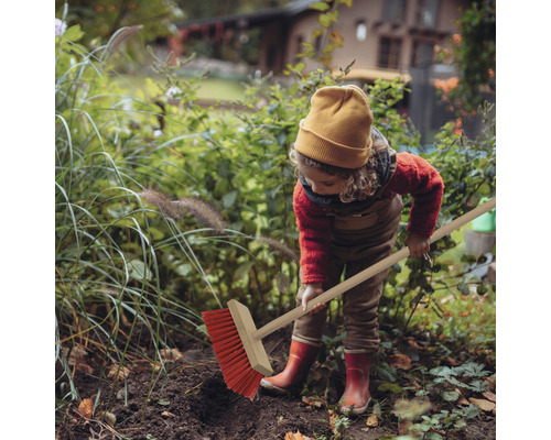 Ein Kind fegt mit einem Straßenbesen im Garten Laub zusammen.