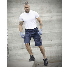 Mann in Shorts, T-Shirt, Handschuhen und Sicherheitsschuhen vor Betonwand.