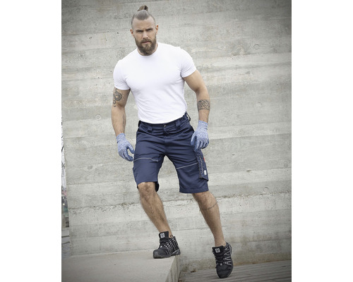 Mann in Shorts, T-Shirt, Handschuhen und Sicherheitsschuhen vor Betonwand.