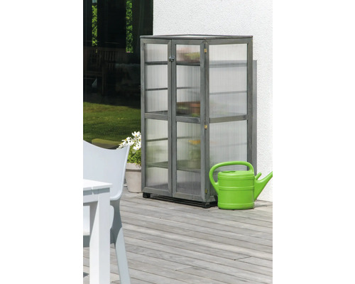 Grauer Holzschrank mit Regalen und durchsichtigen Paneelen steht auf einer Terrasse neben einer grünen Giesskanne.