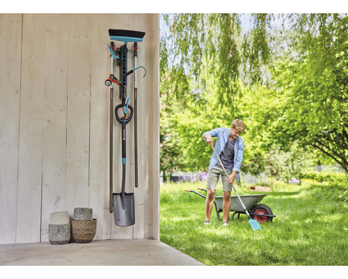 Scène de jardin avec support mural pour outils, bêche, balai, tuyau d''arrosage et lance d''arrosage