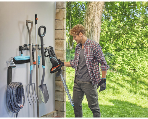 Une personne tenant un taille-haie devant un support mural avec des outils de jardin.