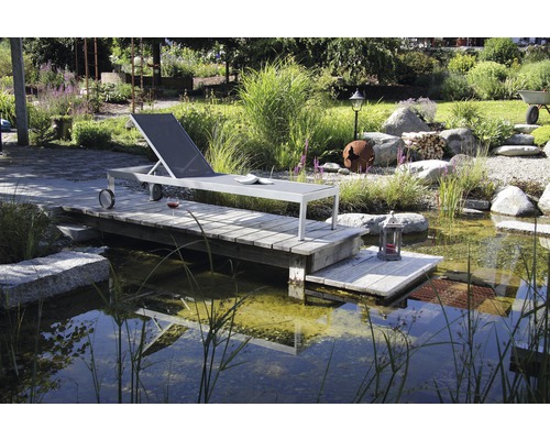 Scène avec une chaise longue en métal sur une plateforme en bois au bord d''un étang dans un jardin.