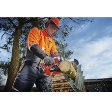 Une personne portant des vêtements de protection coupe un morceau de bois sur un chevalet de sciage avec une tronçonneuse à batterie.