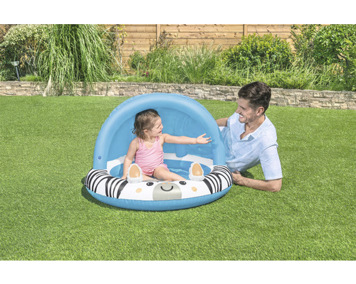 Kind im Planschbecken mit Zebra Design und Sonnenschutz, ein Erwachsener liegt daneben auf dem Rasen