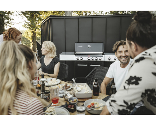 Szene mit einer Gruppe von Personen, die an einem Tisch sitzen, mit Speisen und einem Enders Grill im Hintergrund.