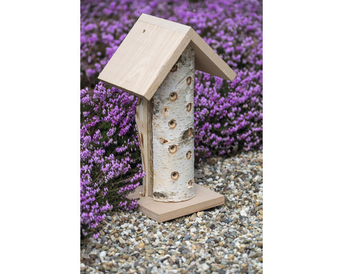 Insektenhotel aus Birkenholz mit Dach und Bohrlöchern vor lila blühenden Pflanzen
