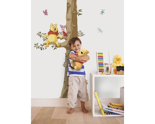 Garçon étreignant une peluche Winnie l''ourson devant un mur avec un autocollant mural Winnie l''ourson et une échelle de croissance.