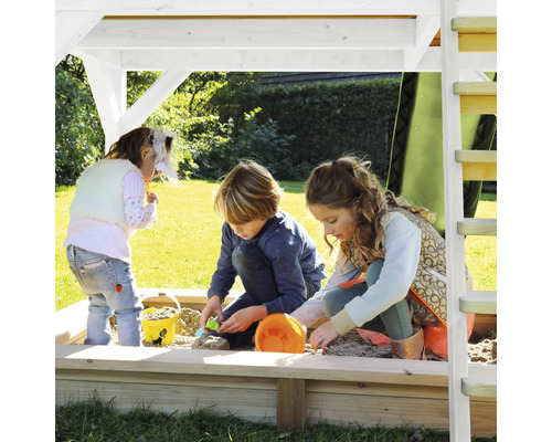Trois enfants jouent dans un bac à sable avec un toit sur une surface verte.