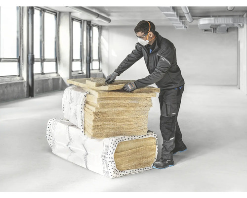 Ein Arbeiter mit Schutzmaske und Schutzbrille schneidet Dämmplatten auf einer Baustelle zu. Verpackte Dämmstoffplatten sind im Hintergrund zu sehen.