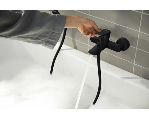 Eine Person bedient eine schwarze Badewannenarmatur, aus der Wasser in eine mit Schaum gefüllte Badewanne läuft.