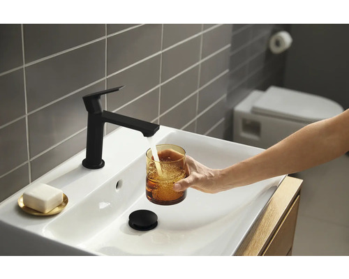 Modernes Badezimmer mit Waschbecken, schwarzer Armatur und einer Person, die ein Glas Wasser füllt.