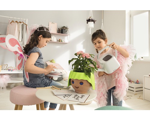 Zwei Mädchen spielen in einem Zimmer mit einer Pflanze in einem Topf mit Gesicht und einer Gießkanne.