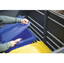 Eine Person öffnet den Deckel einer blauen Mülltonne in einer Mülltonnenbox.