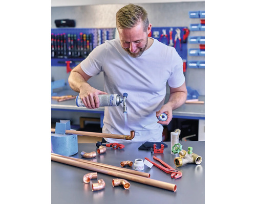 Un homme soude des tuyaux de cuivre avec un chalumeau et de l''étain, entouré d''outils et de tuyaux.