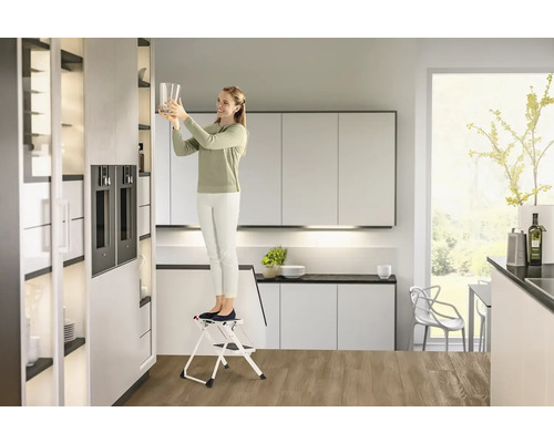 Frau auf einer Trittleiter in einer modernen Küche, die ein Glas in einen Schrank stellt