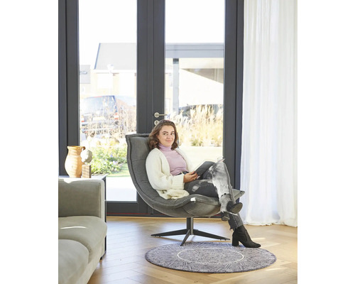 Eine Frau sitzt in einem Sessel vor einer Terrassentür in einem hellen Raum.