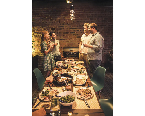 Eine Gruppe von Freunden versammelt sich an einem Tisch im Freien, mit einem Grill im Hintergrund, um gemeinsam zu essen und zu feiern.