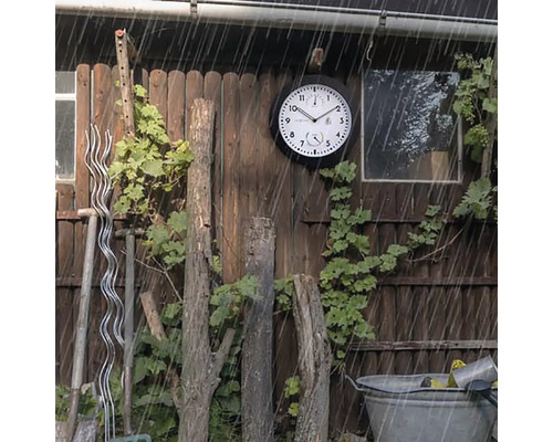 Gartenszene mit einer wetterfesten Außenuhr an einer Holzwand im Regen.