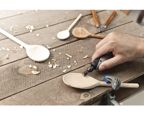 Un homme ponce une cuillère en bois avec un outil multifonction sur une surface en bois. Diverses cuillères en bois et outils de sculpture sont également visibles.