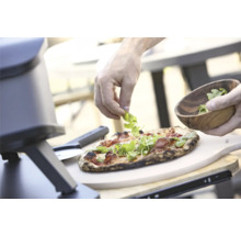 Pizza wird mit Rucola belegt, neben einem Pizzaofen und Pizzaschieber.