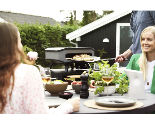 Szene im Freien mit Pizzaofen, Pizza auf Pizzaschieber und Personen am Tisch
