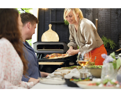 Scène avec des personnes, four à pizza et pizza servie sur une planche à pizza.