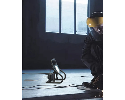 Arbeiter mit Schutzvisier arbeitet mit einem tragbaren LED-Strahler auf einer Baustelle.