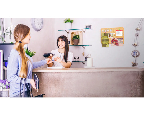 Eine Kundin bezahlt an der Kasse mit Kreditkarte, im Hintergrund eine Kork-Pinnwand