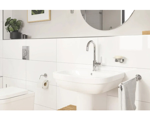 Salle de bain lumineuse avec lavabo, robinet, miroir et toilettes devant des murs carrelés blancs.