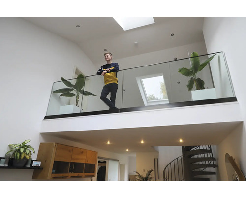 Un espace de vie lumineux avec galerie, garde-corps en verre, plantes d''intérieur et une personne.