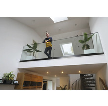 Ein junger Mann steht auf einer Galerie mit Glasgeländer in einem hellen, modernen Wohnraum mit Zimmerpflanzen und Oberlichtern.